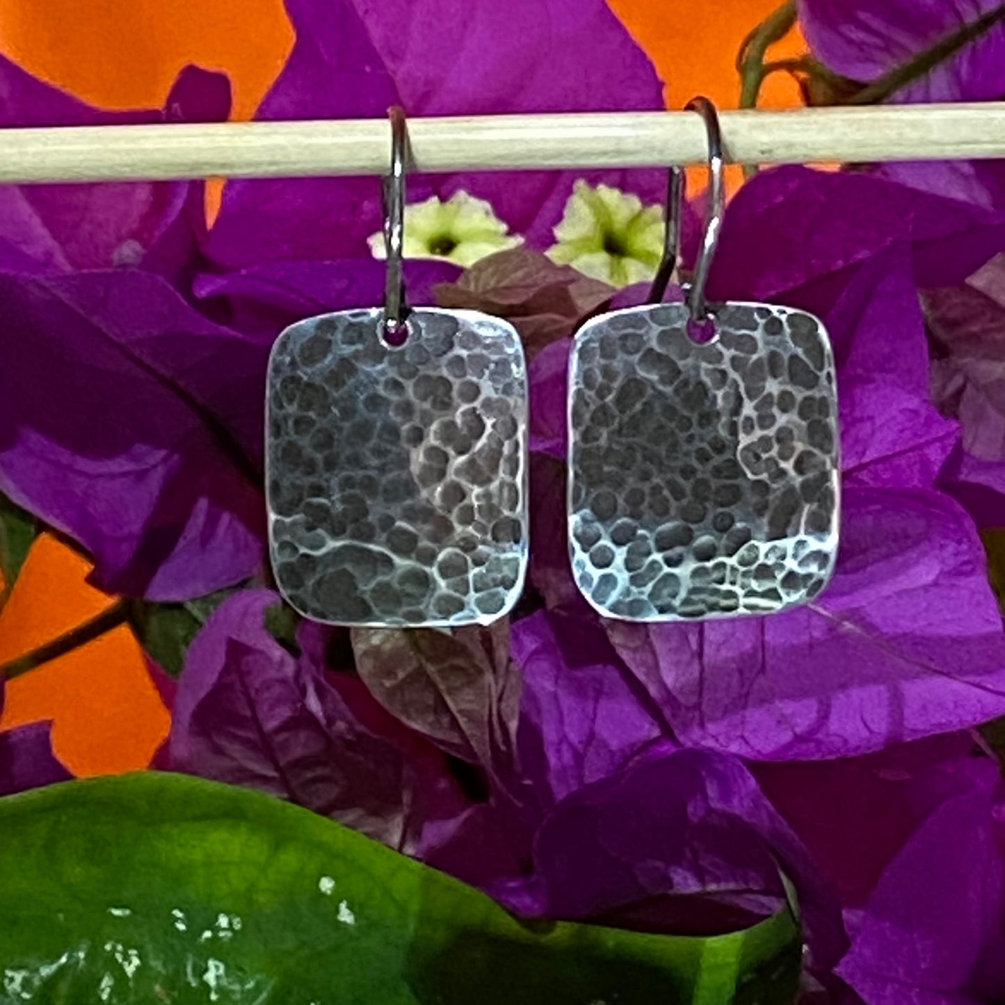 Aretes martillados y pavonados colgando de bambú sobre buganvilla violeta, con fondo naranja. Amuleto Artesanal de Plata Fina 950 (Tunja).