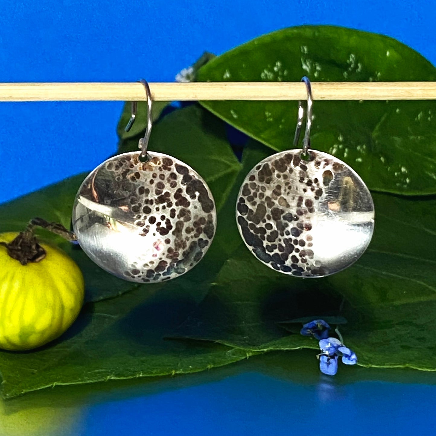 Par de Aretes de Plata Fina 950 (Plata 925), con el fruto silvestre amarillo más visible. La composición incluye hojas de monte sobre fondo azul, destacando el acabado martillado.