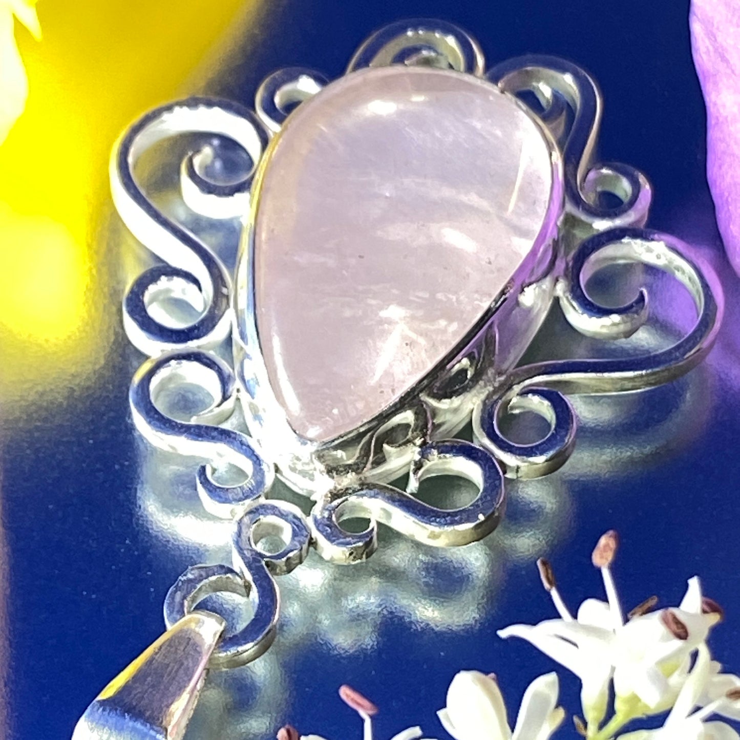 Dije de Plata 950 con Joyas con Historia. Colgante de Cuarzo Rosado con engaste de volutas. Fondo azul metálico con hojas flores blancas, amarillas y violetas alrededor de la pieza. Vista superior. Primer plano. Mucho brillo y mucha luz en la foto.