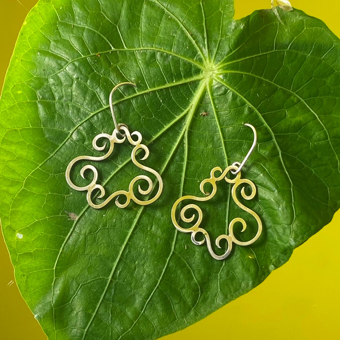 Aretes de Plata Fina 950 de diseño calado. La pieza está montada en una hoja tropical grande cuya transparencia es resaltada por la luz fuerte sobre fondo amarillo (Dos Quebradas).