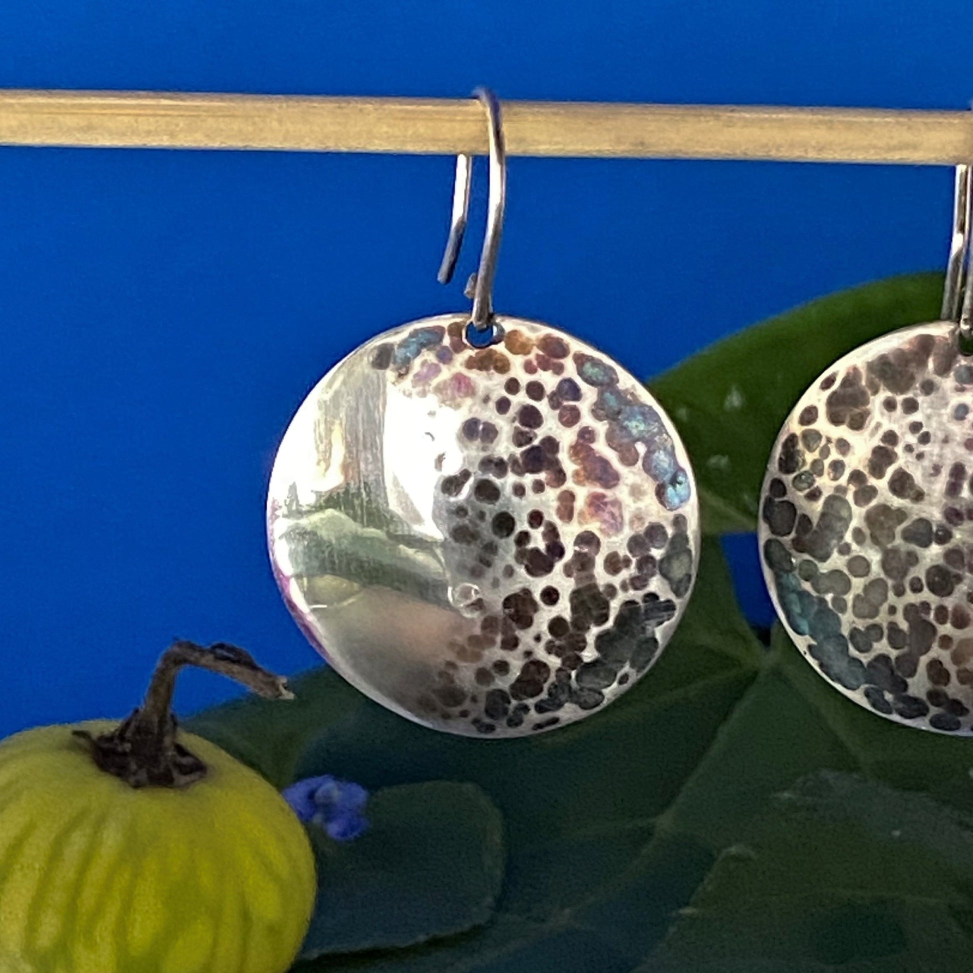 Aretes circulares de Plata Fina 950 (martillados y pavonados) con Materiales Naturales. El diseño de media luna se complementa con la decoración de fruta y hojas silvestres sobre un fondo azul contrastante.