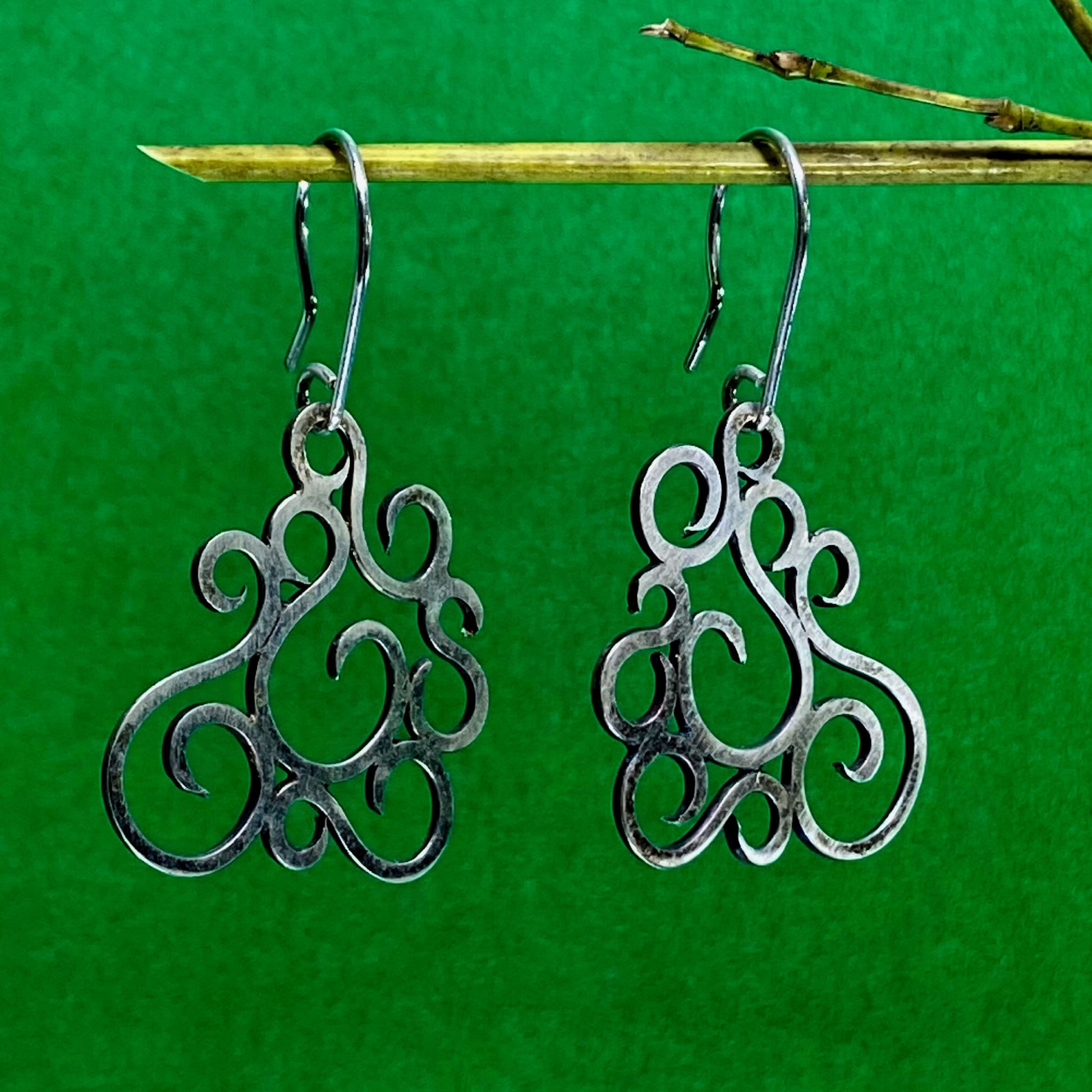 Aretes de Plata Fina 950 con Arte Colombiano, diseño calado de volutas suspendidos en bambú, fondo verde.