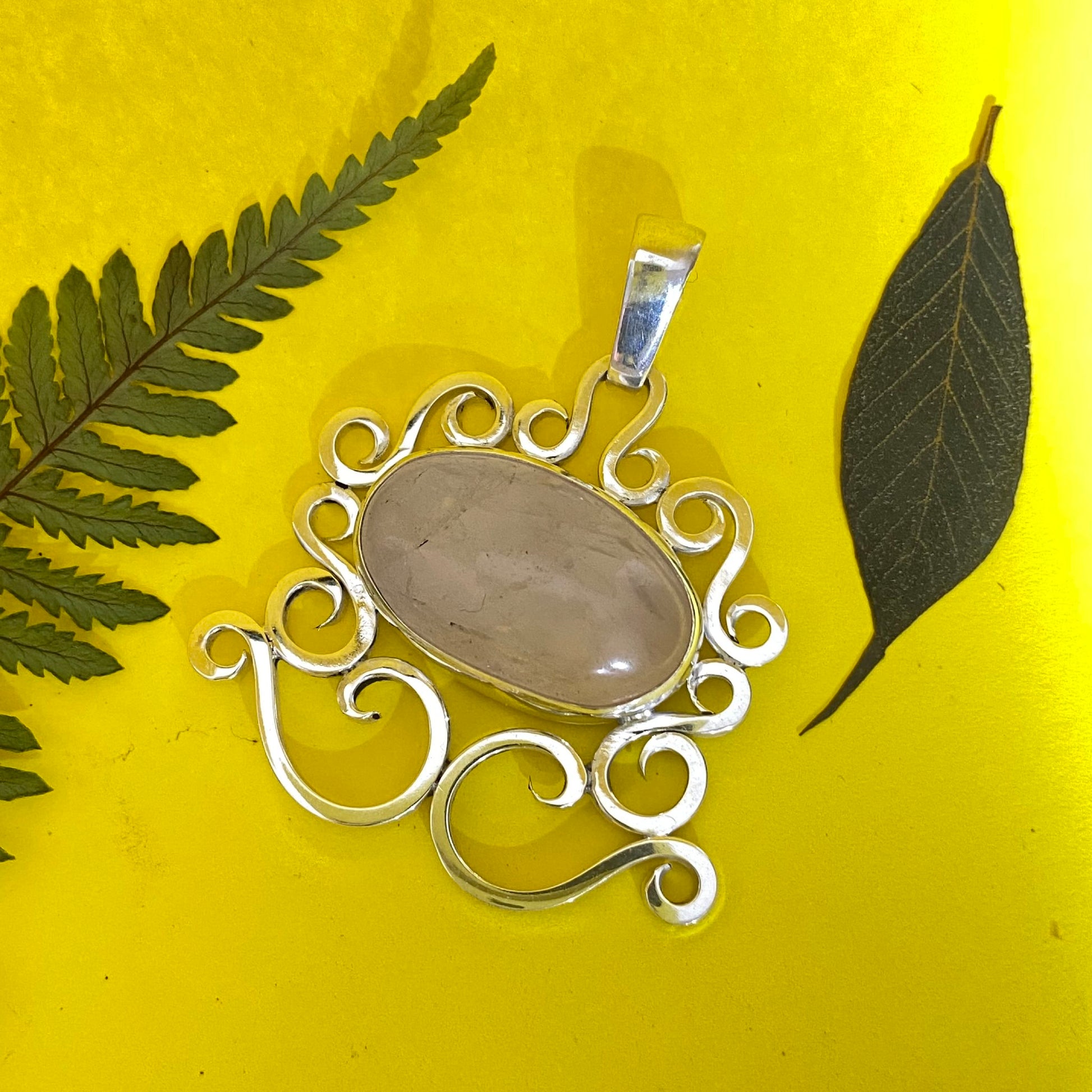 Dije de Plata Fina 950 con Diseño Orgánico de volutas y Cuarzo Rosa ovalado. Sobre fondo amarillo brillante, decorado con hojas de helecho y hoja oscura (Colombia).