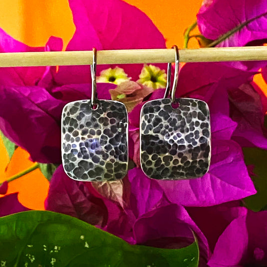 Par de Aretes Hechos a Mano en Plata Fina 950. Diseño rectangular martillado y pavonado (oscurecido), colgado sobre un fondo de flores buganvillas violeta y hojas verdes.