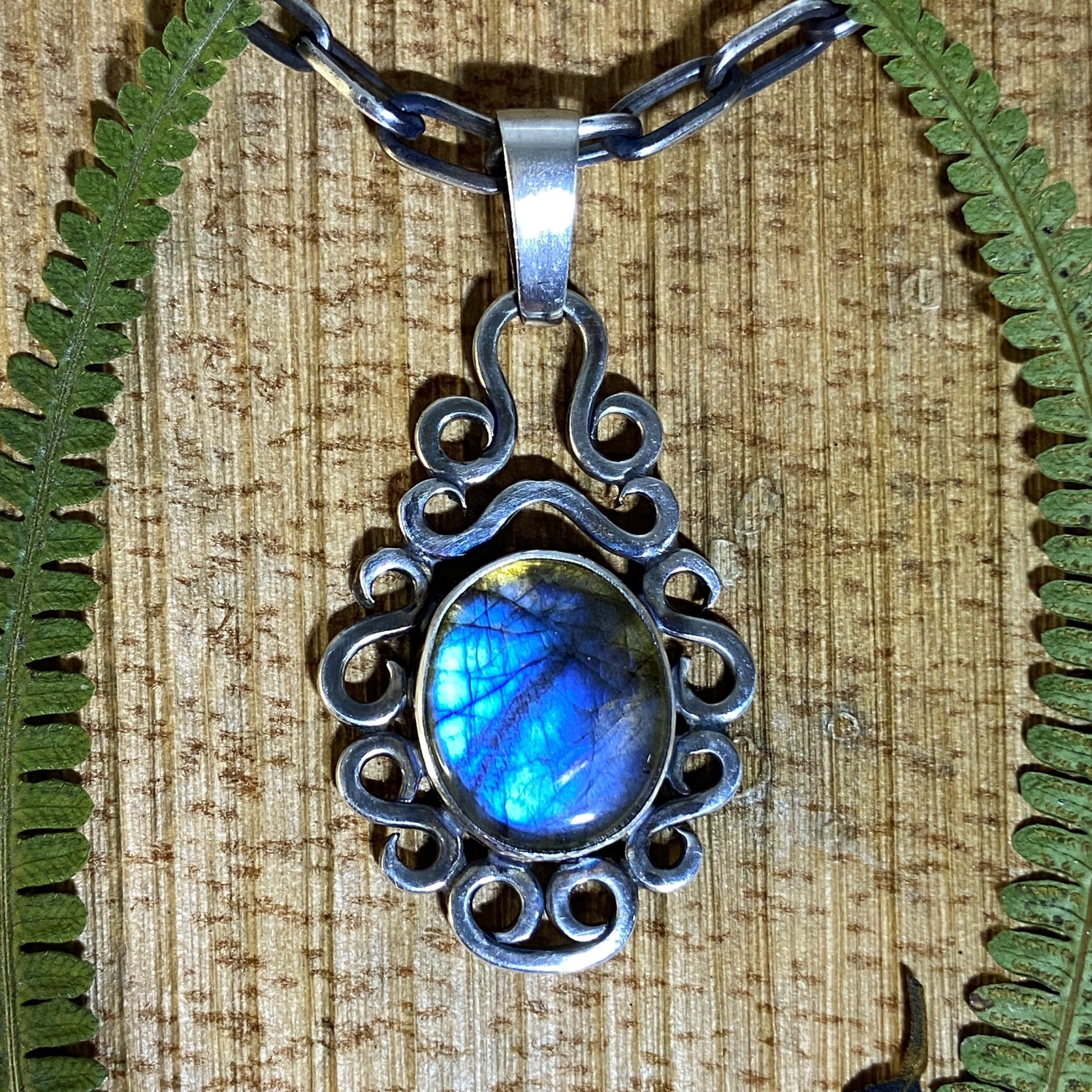 Dije de Plata Fina 950 con Diseño Rústico (Colombia). Colgante de Labradorita iridiscente en engaste de plata con volutas, sobre fondo de madera natural y enmarcado con hojas de helecho.