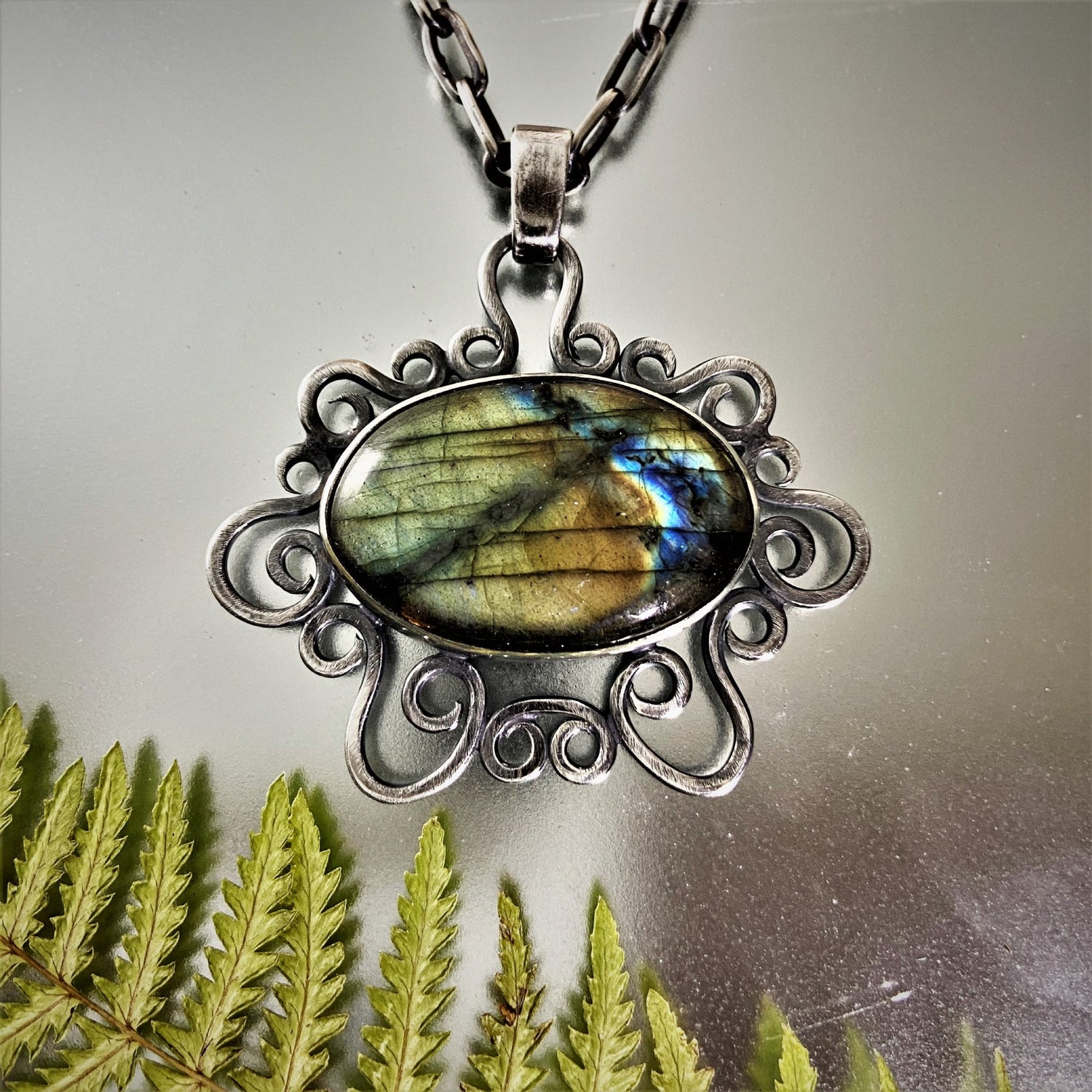 Dije de Plata Fina 950 (Santa Marta). Colgante de Labradorita con engaste de volutas. Foto sobre fondo metálico reflectante. Decorado con hojas de helecho en la parte inferior.