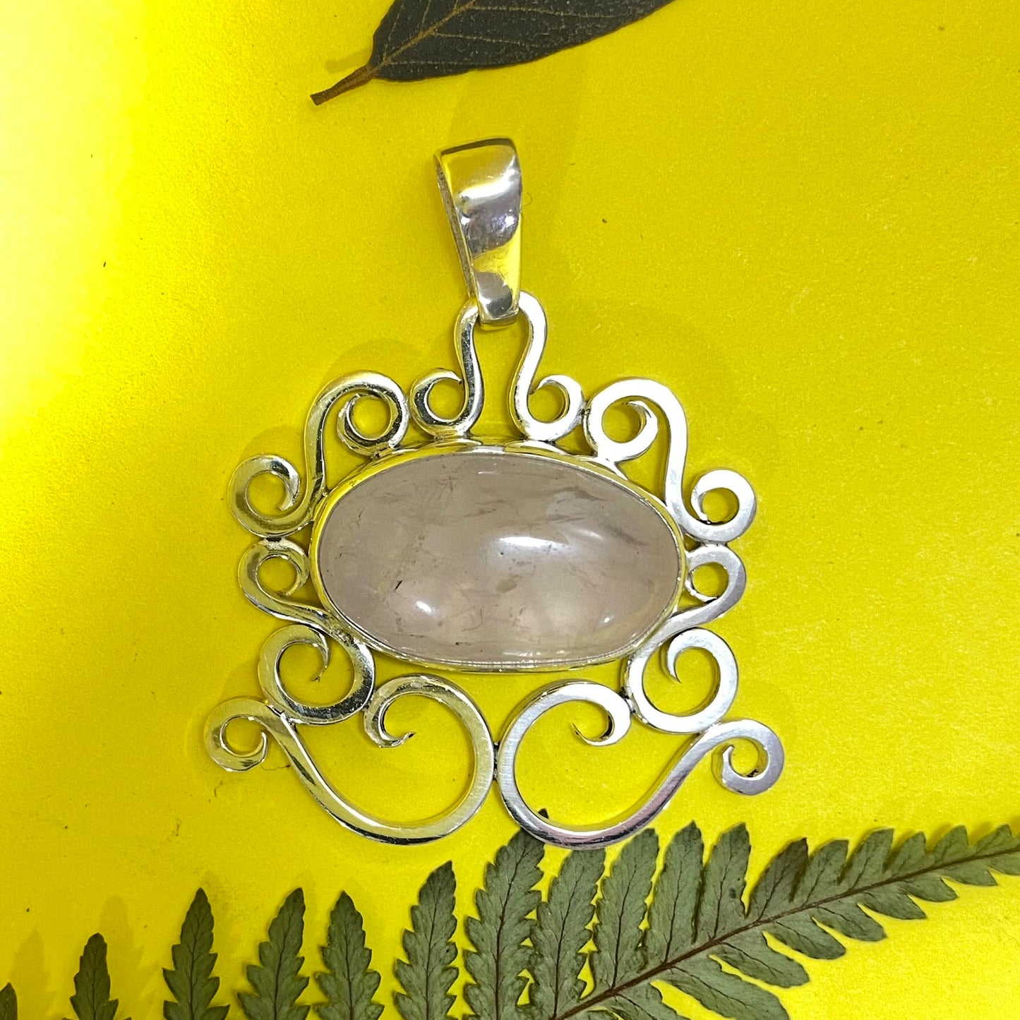 Dije de Colección Limitada en Plata 950. Primer plano del Cuarzo Rosa ovalado, con fondo amarillo. La pieza está enmarcada por hojas de helecho (Apartadó).