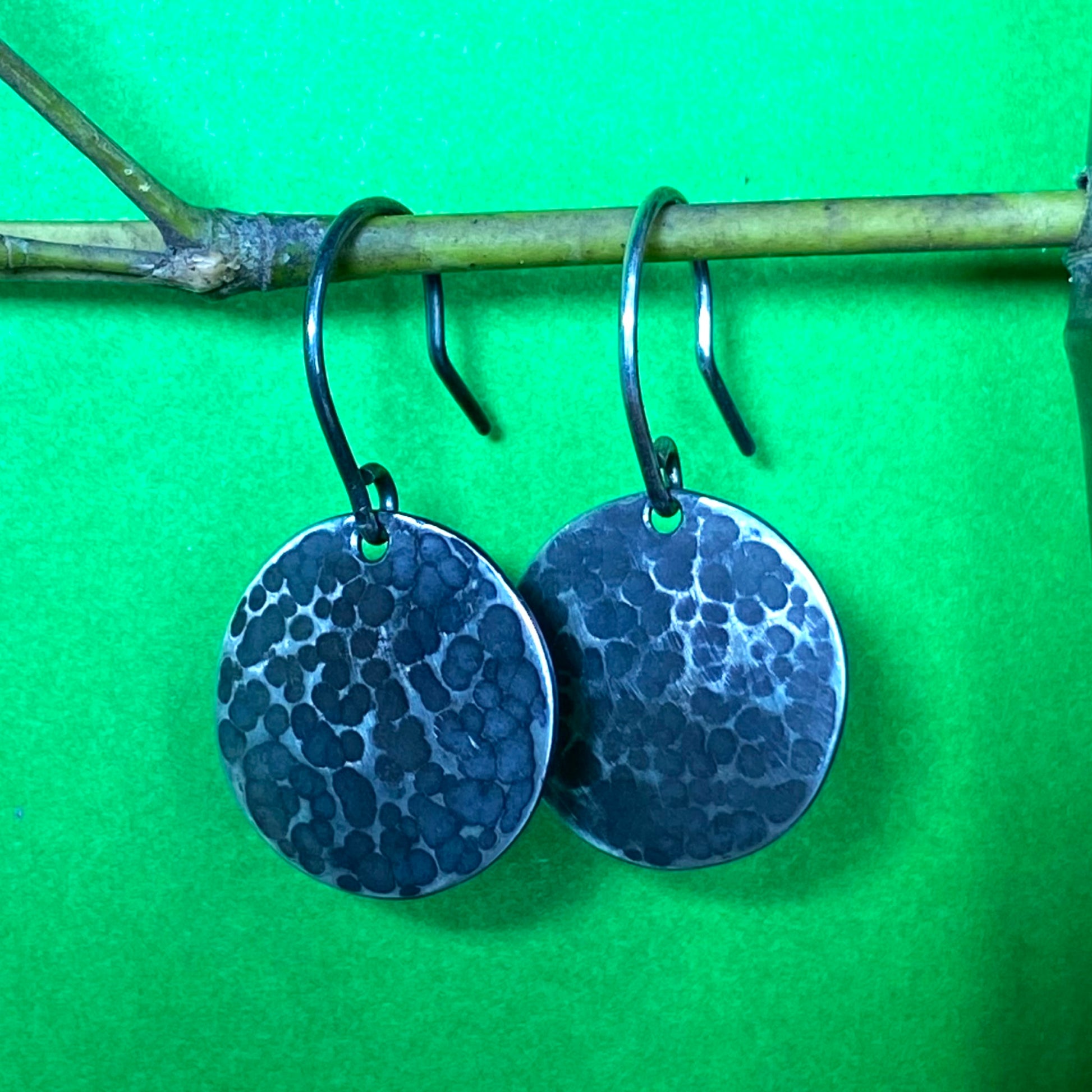 Aretes de Plata Fina 950 de Diseño Minimalista. Par de joyas martilladas y pavonadas colgando de bambú, con luz intensa y sombras sobre fondo verde (Ibagué).