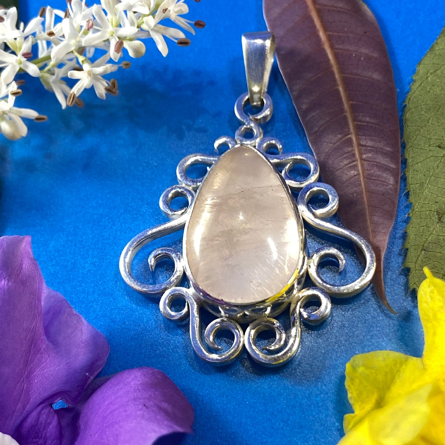 Dije de Plata Fina 950. Colgante con cuarzo rosado sobre fondo azul intenso. Decoración con flores purpura, flores blancas, hojas seca, flores amarillas,