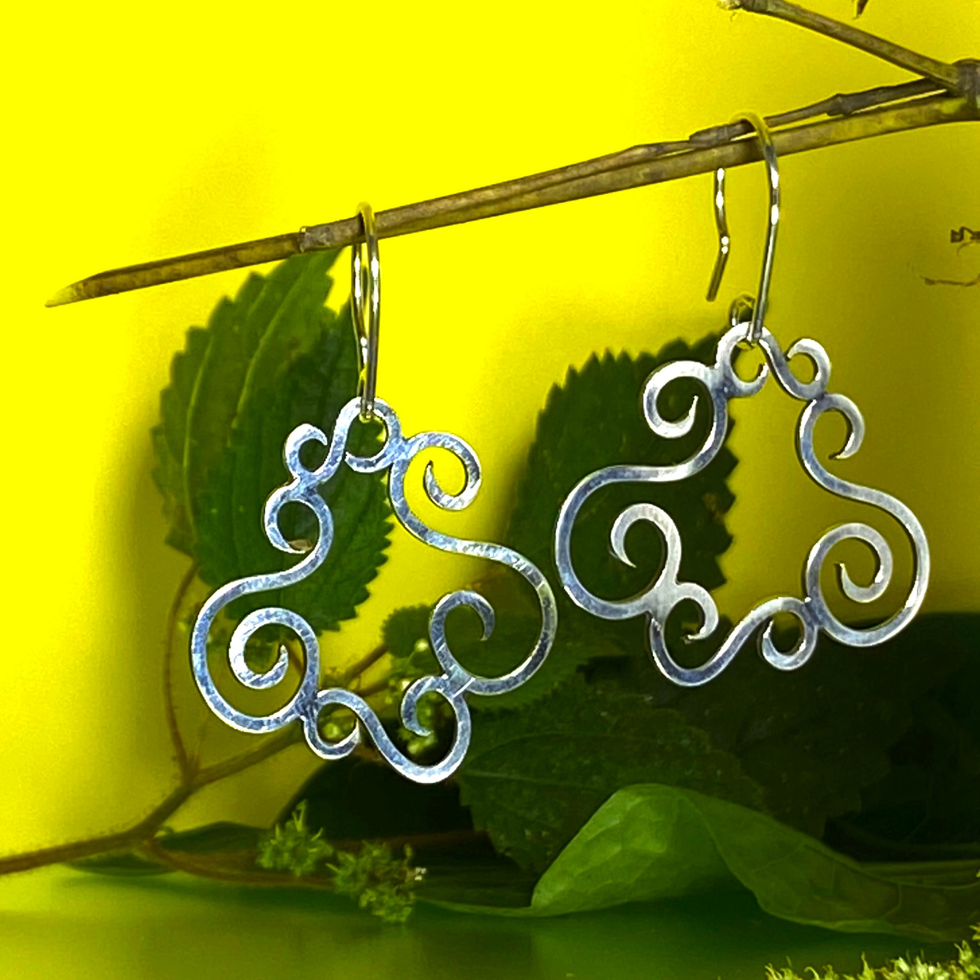 Aretes de Plata Fina 950 de Estilo Bohemio, con hojas de monte como decoración. Vistos sobre una superficie semireflectiva con fondo amarillo vibrante.