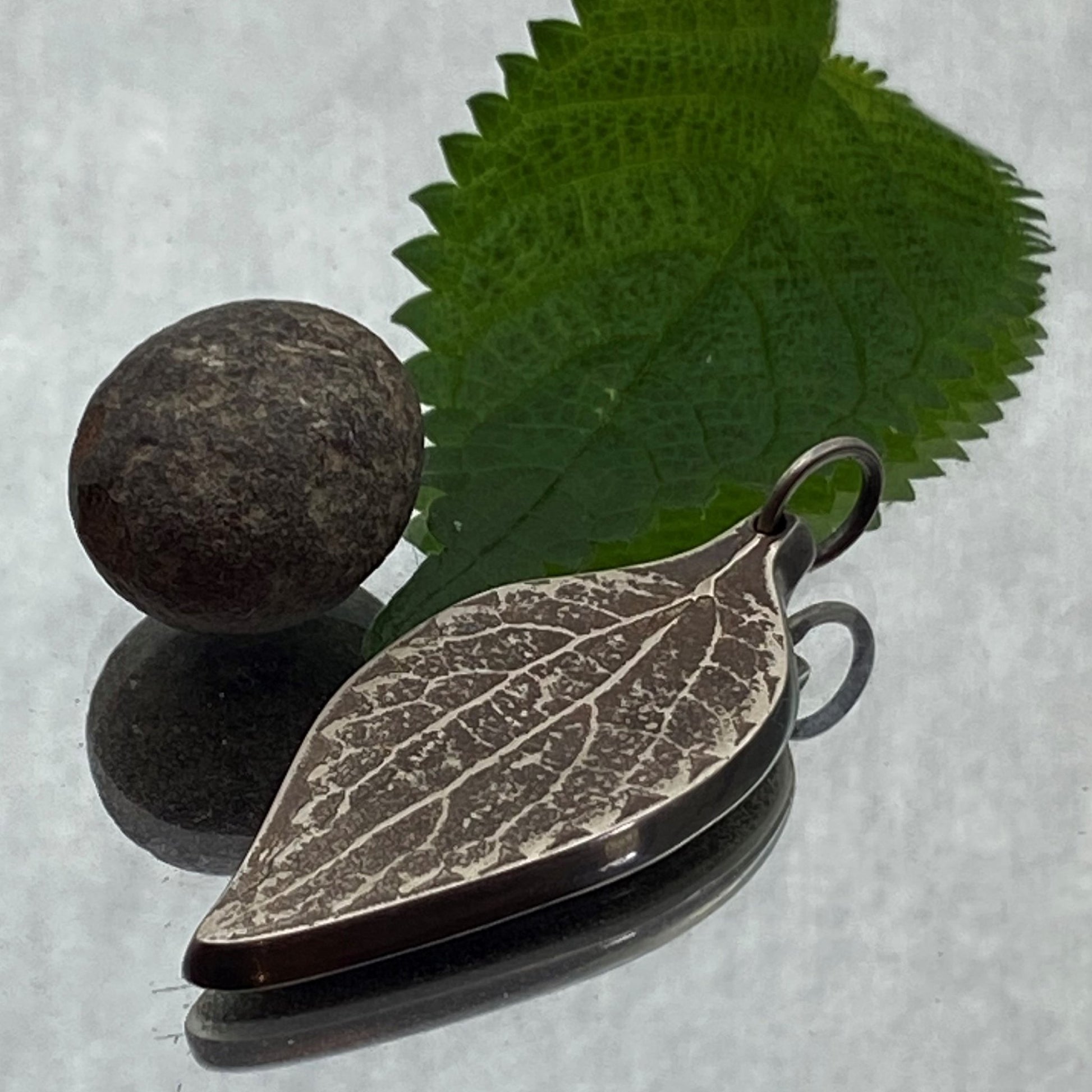 Dije de Plata Fina 950 con Inspiración Ancestral, diseño de hoja martillada y oxidada. La pieza descansa sobre una superficie reflectante con una piedra redonda oscura y una hoja verde de fondo (Dos Quebradas).