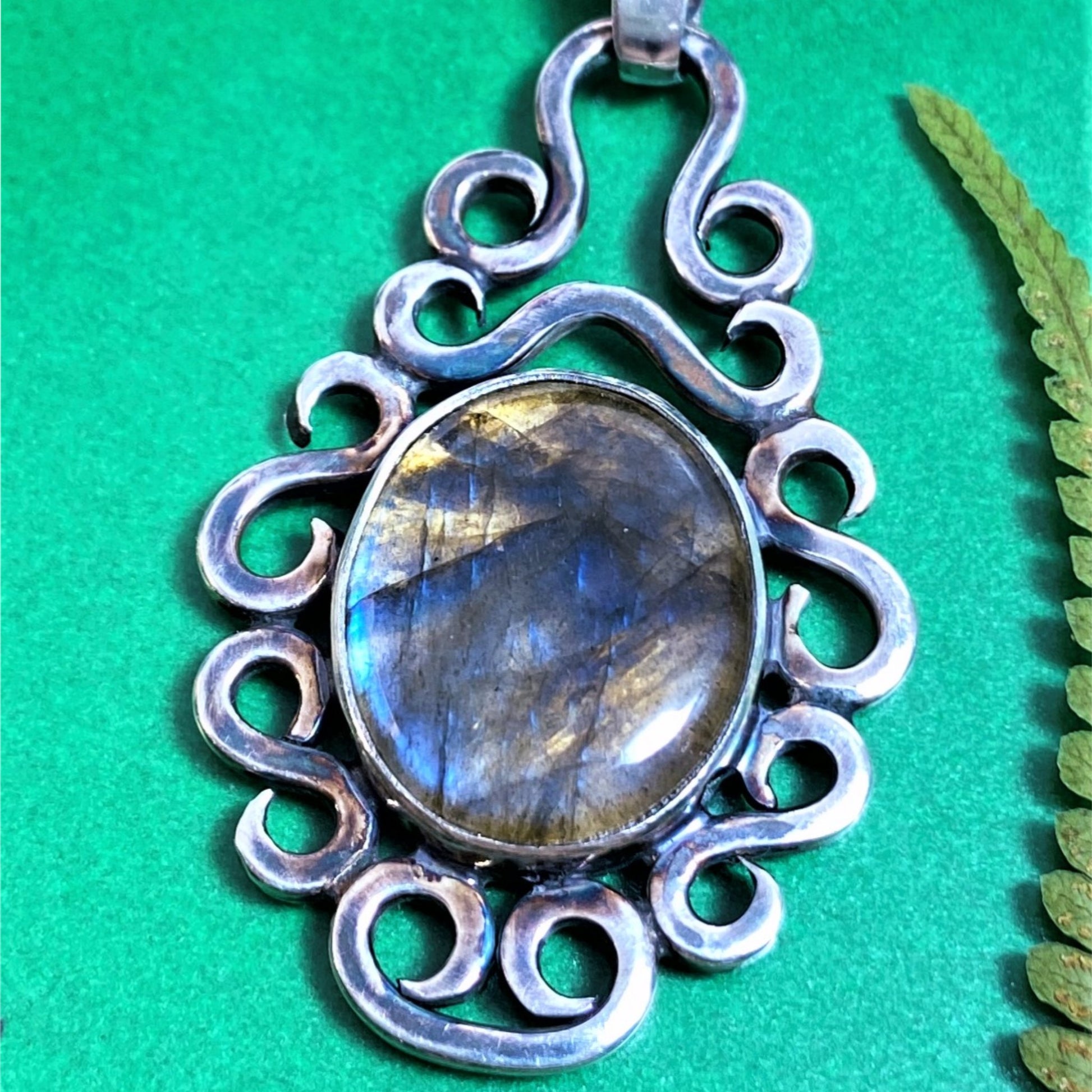 Dije de Plata Fina 950 con Diseño con Significado. Primer plano de la Labradorita ovalada en un engaste de plata con volutas sobre un fondo verde brillante (Florencia).