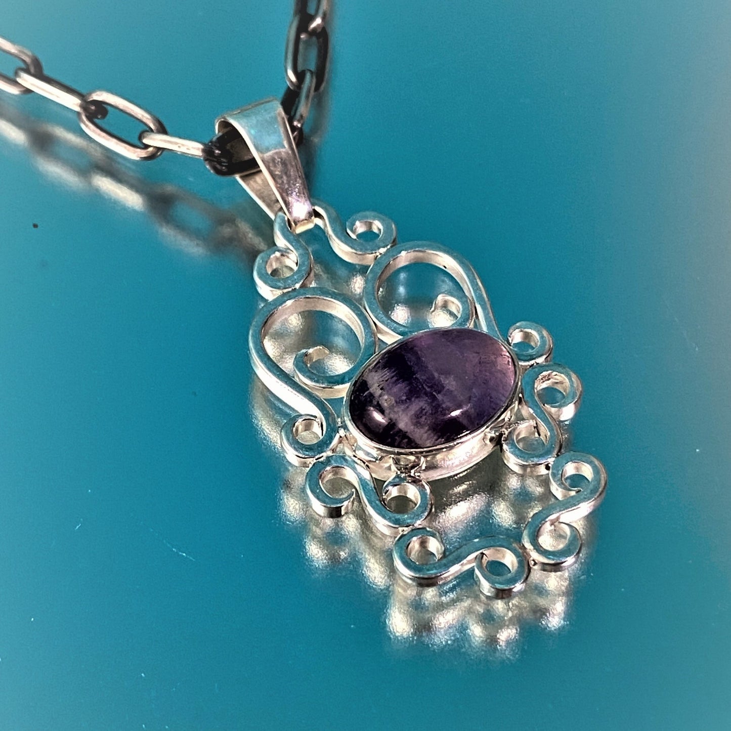 Dije de Plata Fina 950 con Estilo Minimalista Elegante. Gema Amatista ovalada, presentada en ángulo sobre un fondo azul metálico reflectante (Girardot).