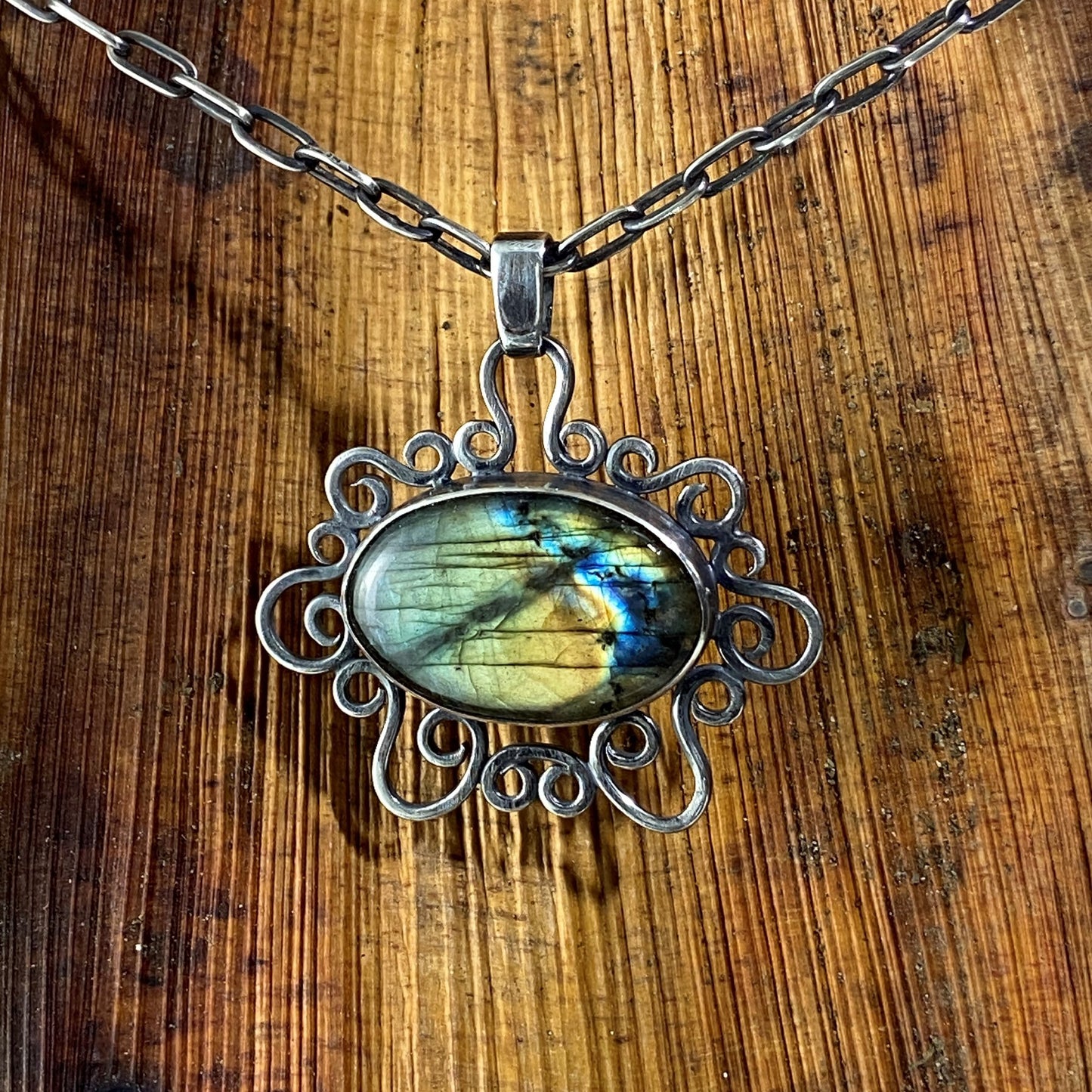 Dije de Plata 950, Plata Pura. La gema Labradorita (tonos verde/azul/amarillo) cuelga de una cadena pavonada, sobre fondo de madera natural rústica (Ocaña).