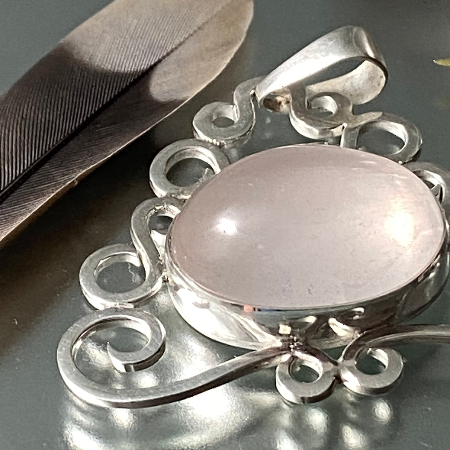 Pieza Exclusiva en Plata Fina 950. Dije de Cuarzo Rosado ovalado en primer plano, decorado con pluma y sobre un fondo reflectante gris (Rionegro).
