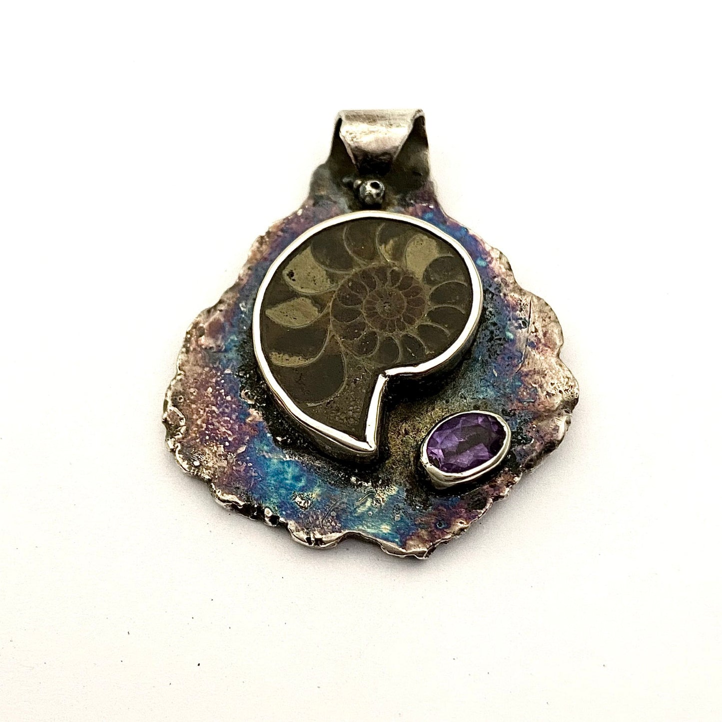 Dije de Plata Fina 950 con Inspiración Ancestral, diseño de hoja martillada y oxidada. La pieza descansa sobre una superficie reflectante con una piedra redonda oscura y una hoja verde de fondo (Dos Quebradas).