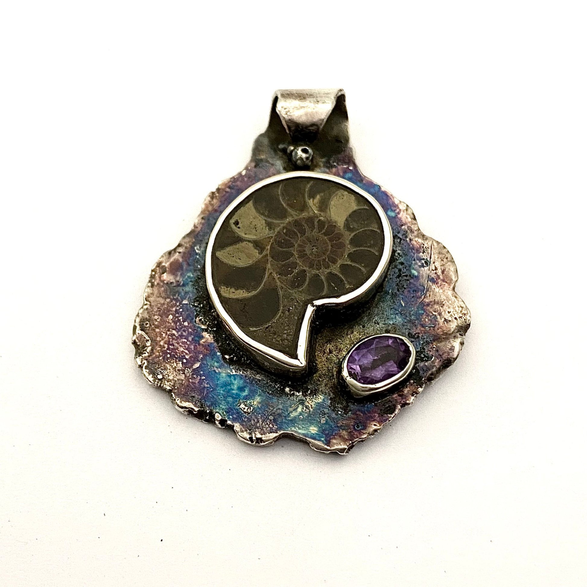 Dije de Plata Fina 950 con Inspiración Ancestral, diseño de hoja martillada y oxidada. La pieza descansa sobre una superficie reflectante con una piedra redonda oscura y una hoja verde de fondo (Dos Quebradas).