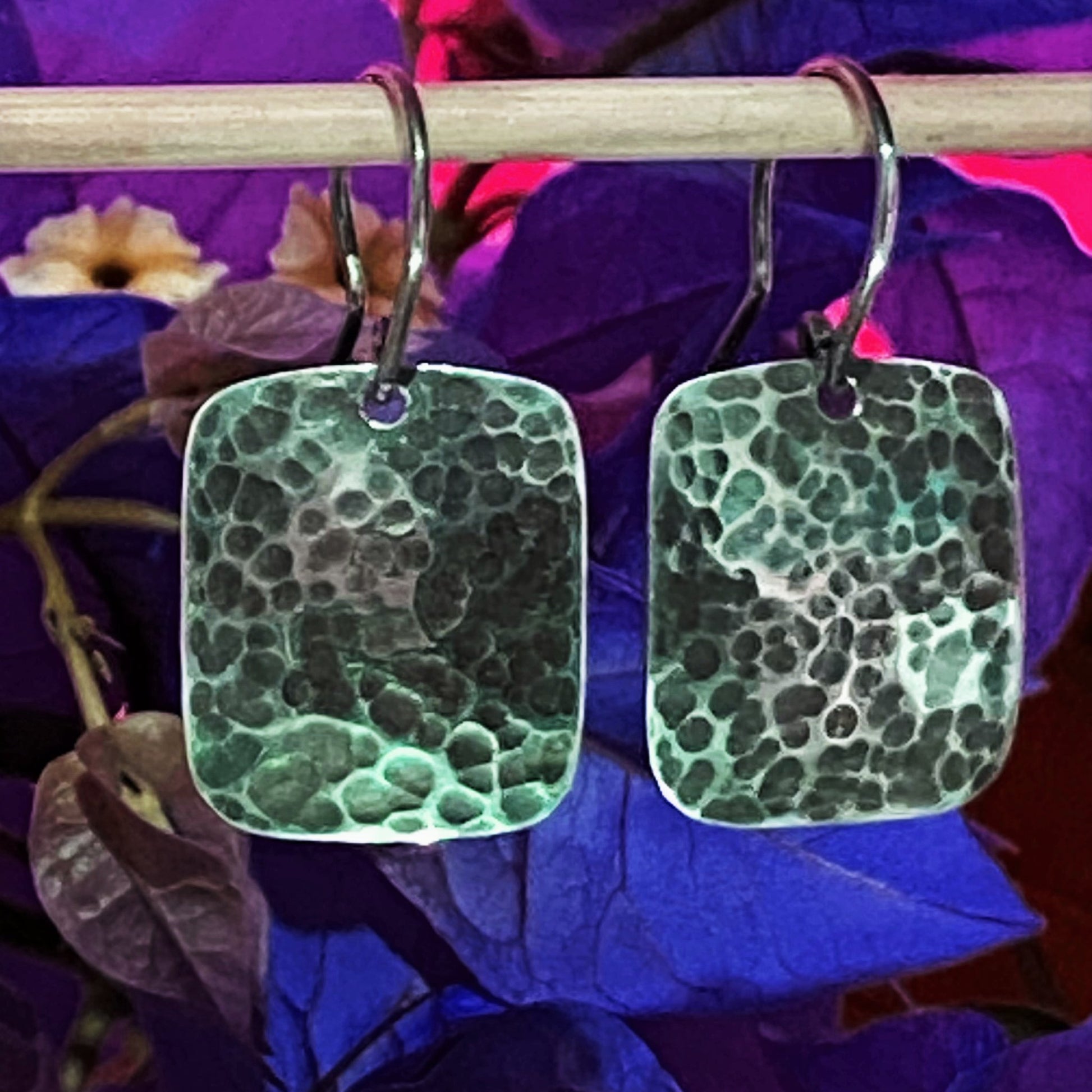 Aretes de Plata 950 con Diseño Colombiano. Primer plano del par rectangular martillado, destacando el acabado pavonado sobre un fondo de buganvilla azul intenso (Tunja).