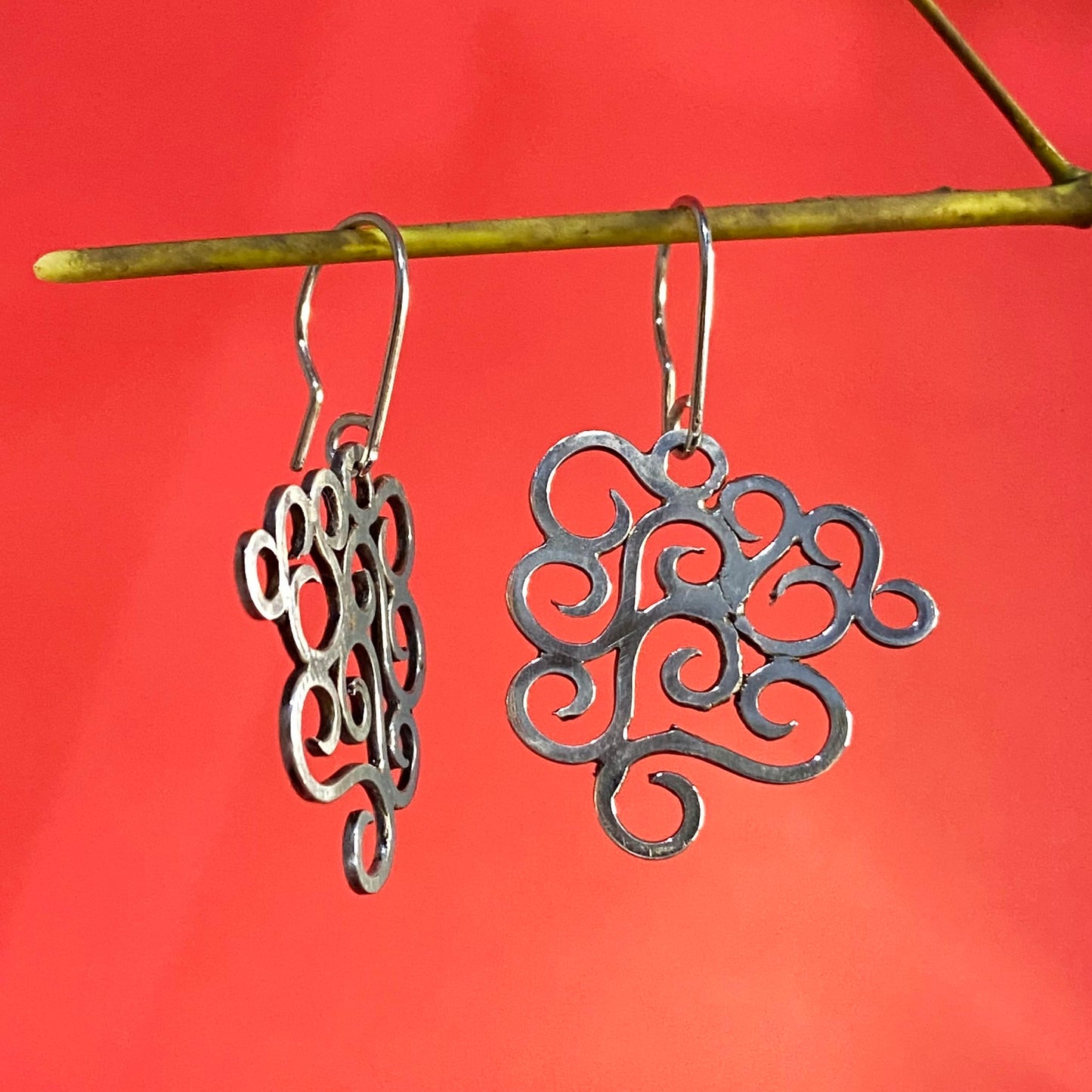 Joyas Hechas en Colombia: Aretes de Plata Fina 950, mostrados de lado para apreciar el grosor del material, colgando de una rama fina con fondo rojo (Valledupar).