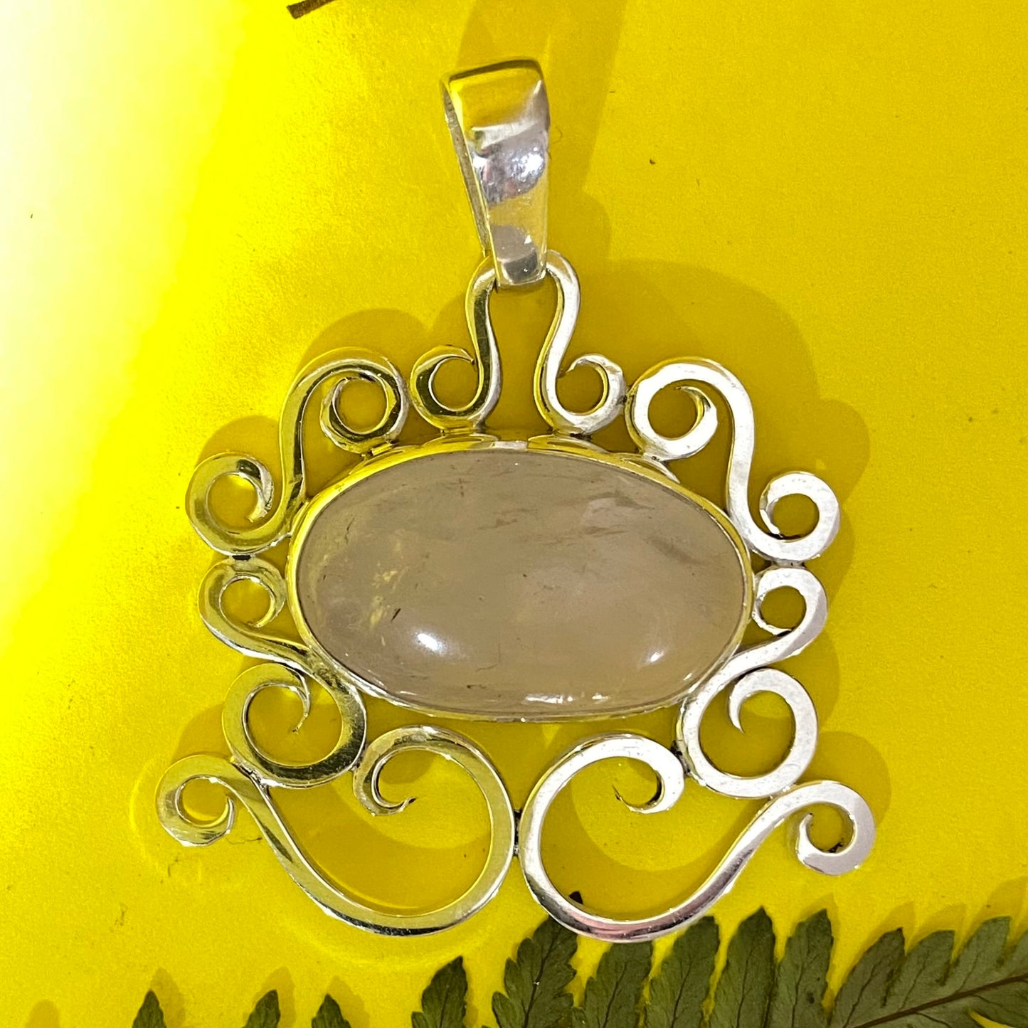 Dije con Gemas Auténticas (Cuarzo Rosa) en Plata Fina 950. La joya con marco de volutas es fotografiada sobre fondo amarillo, con reflejos que resaltan el brillo de la gema (Bogotá).