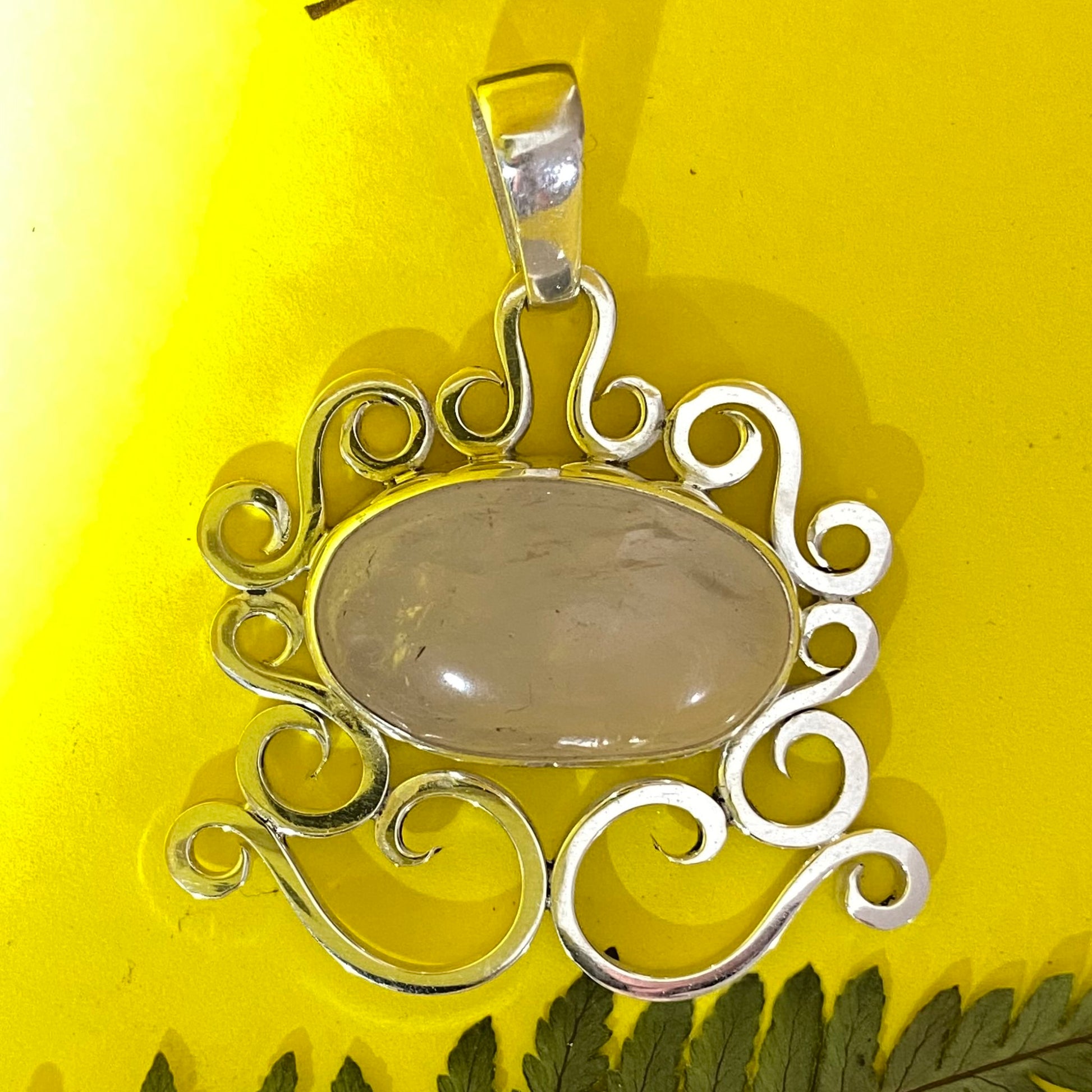 Dije con Gemas Auténticas (Cuarzo Rosa) en Plata Fina 950. La joya con marco de volutas es fotografiada sobre fondo amarillo, con reflejos que resaltan el brillo de la gema (Bogotá).