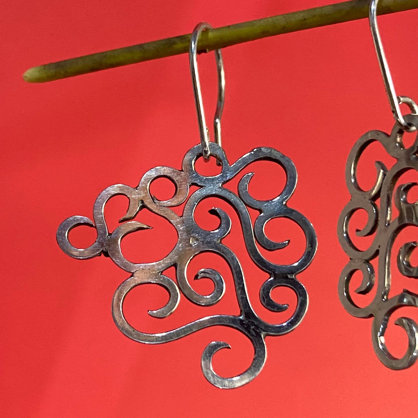  Aretes de Plata Fina 950 con Arte Colombiano, diseño orgánico calado en primer plano sobre fondo rojo vibrante, colgando de una rama.