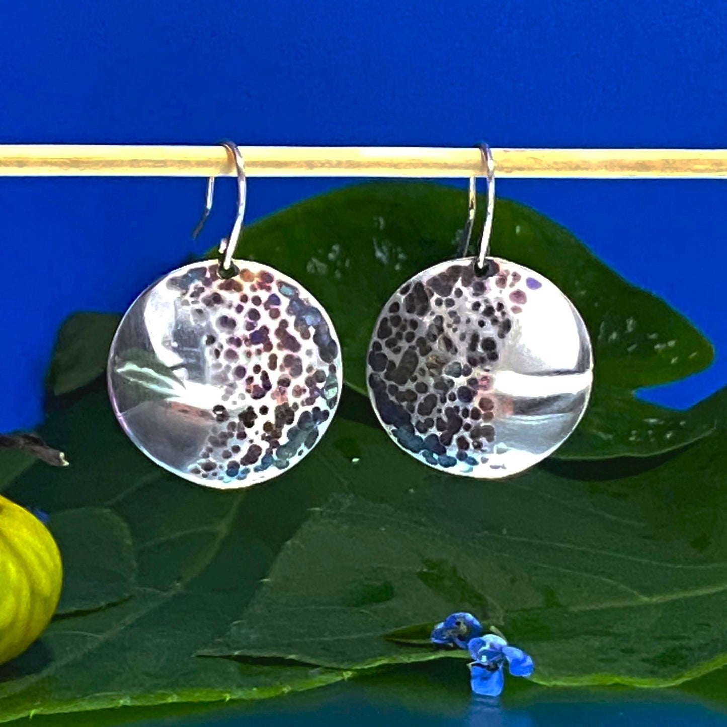 Par de Aretes de Plata Fina 950 de Alta Calidad. Muestra la joya colgando del bambú con un fragmento de fruta silvestre, enfatizando el diseño circular martillado sobre fondo azul.