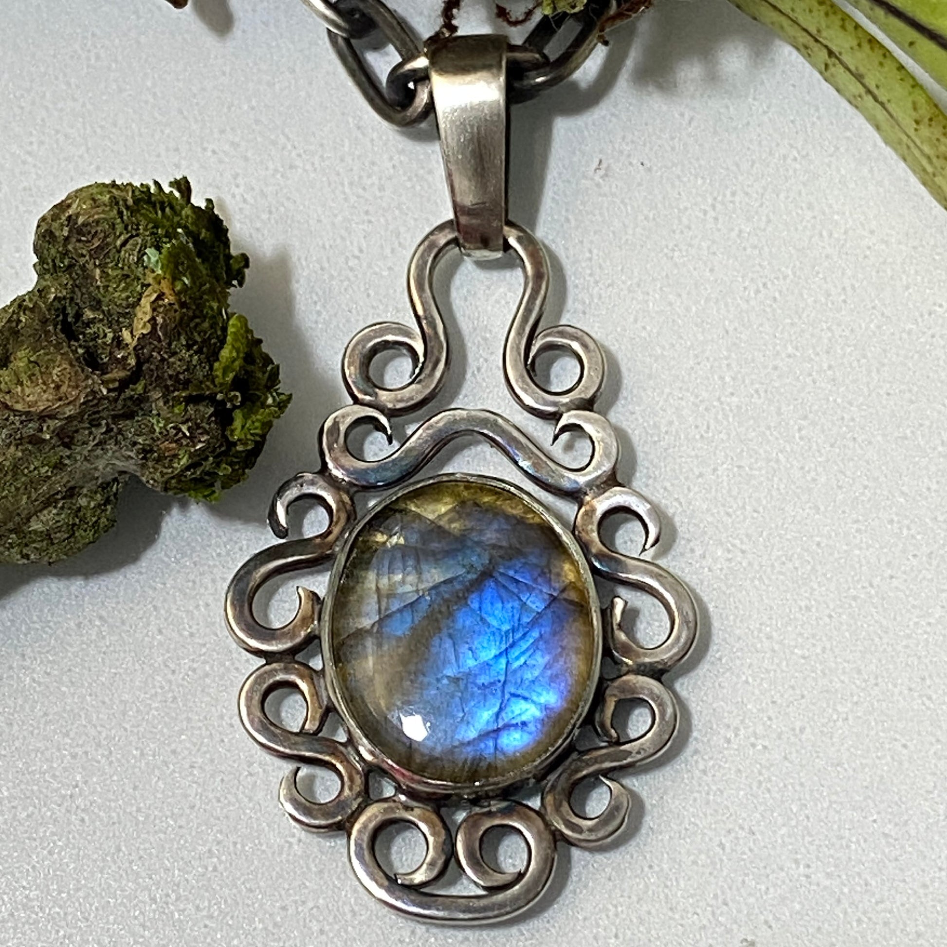Dije de Plata Fina 950, Artesanal. Primer plano de la Labradorita ovalada (brillo azul intenso) en engaste de volutas, con decoración de musgo y cadena gruesa.