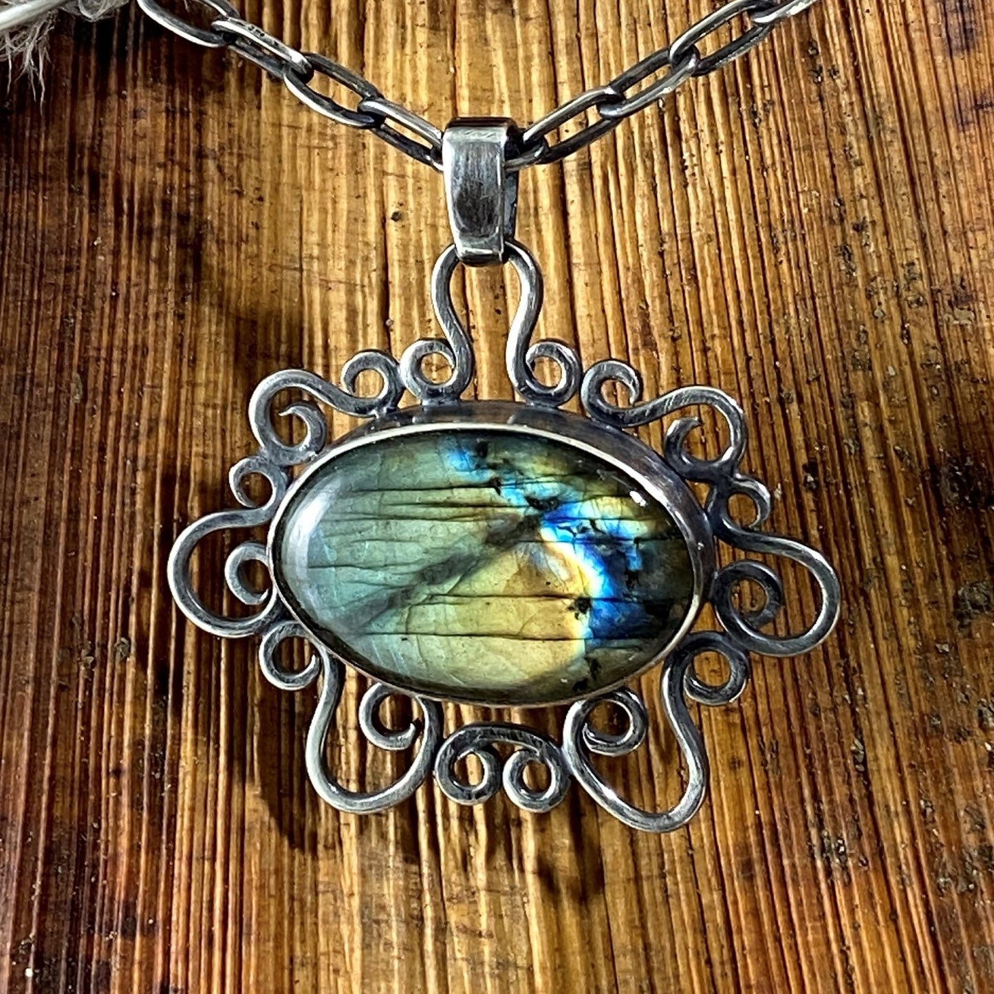 Dije con Cristales Naturales (Labradorita) y marco de volutas. Vista de cerca sobre un fondo de madera, colgando de una cadena pavonada gruesa (Colombia).