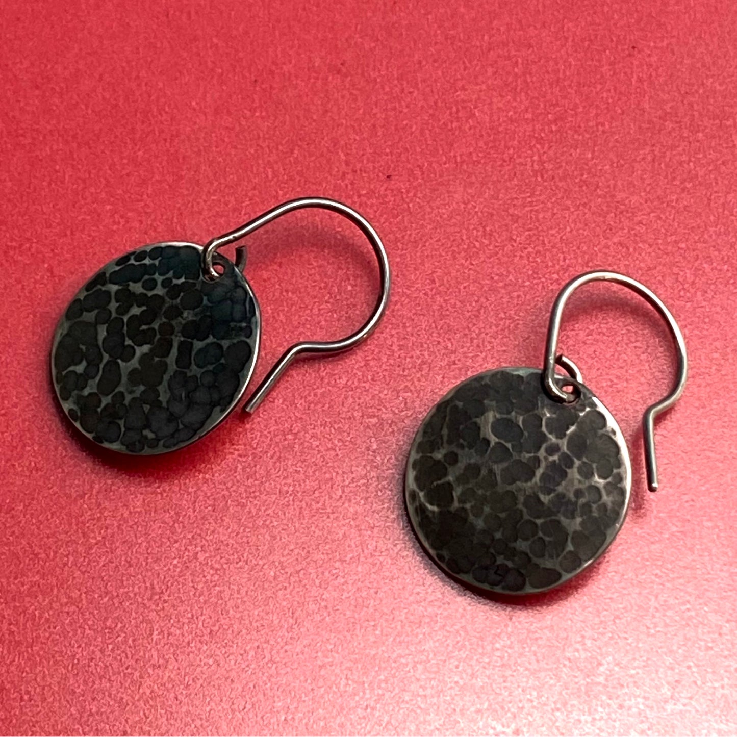 Aretes de Plata Fina 950 con Diseño Orgánico Martillado, colocados sobre un fondo rojo semi mate. El brillo en la parte inferior derecha resalta la textura.