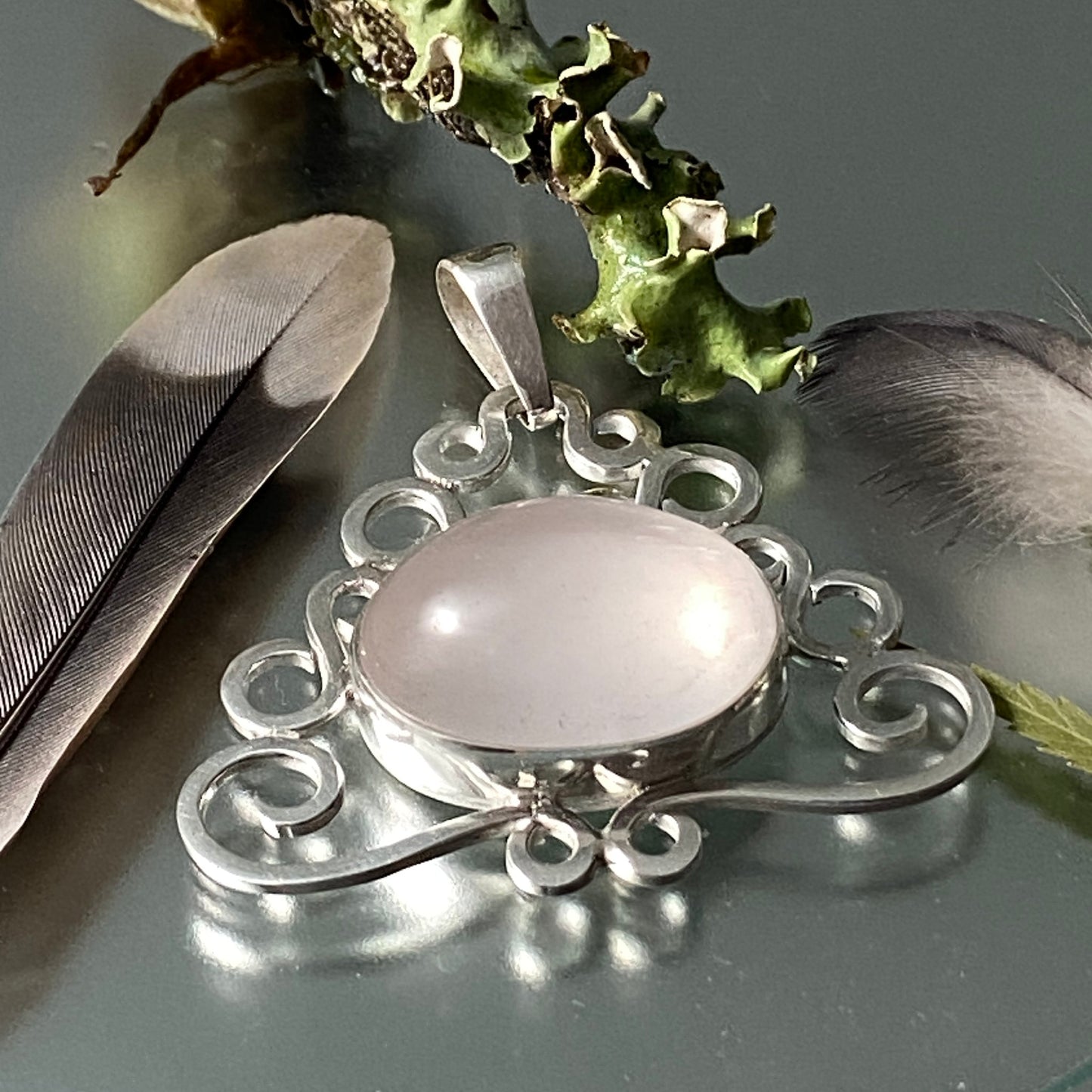 Dije de Piedras Naturales (Cuarzo Rosado) en Plata 950. Composición artística con plumas y liquen/musgo sobre superficie reflectante.