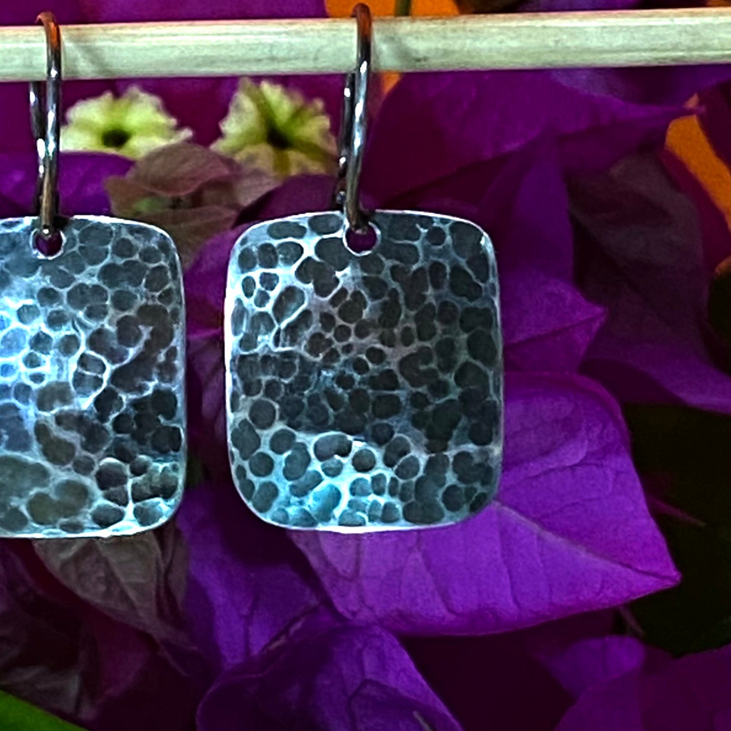 Aretes de Plata 950 con Diseño Colombiano. Primer plano del par rectangular martillado, destacando el acabado pavonado sobre un fondo de buganvilla azul intenso (Tunja).