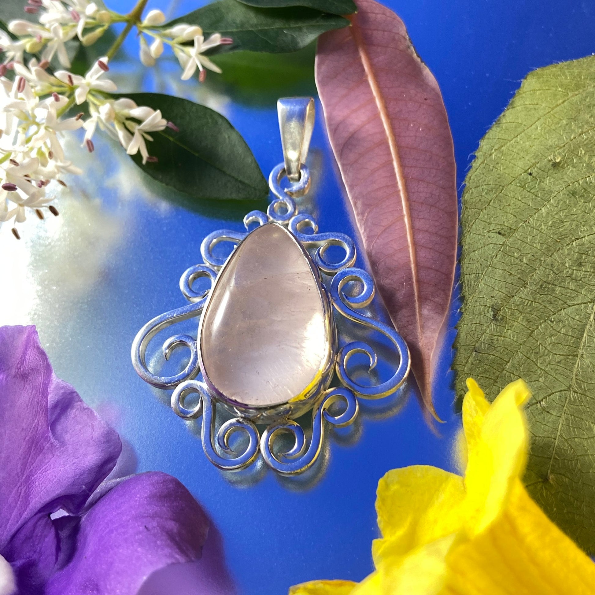 Dije de cuarzo rosado en Plata 950. Sobre superficie azul brillante muy reflectiva. Decorado con flores violetas amarillas y blancas. Mucha luz y un intenso brillo que le da mucha vida a la foto.