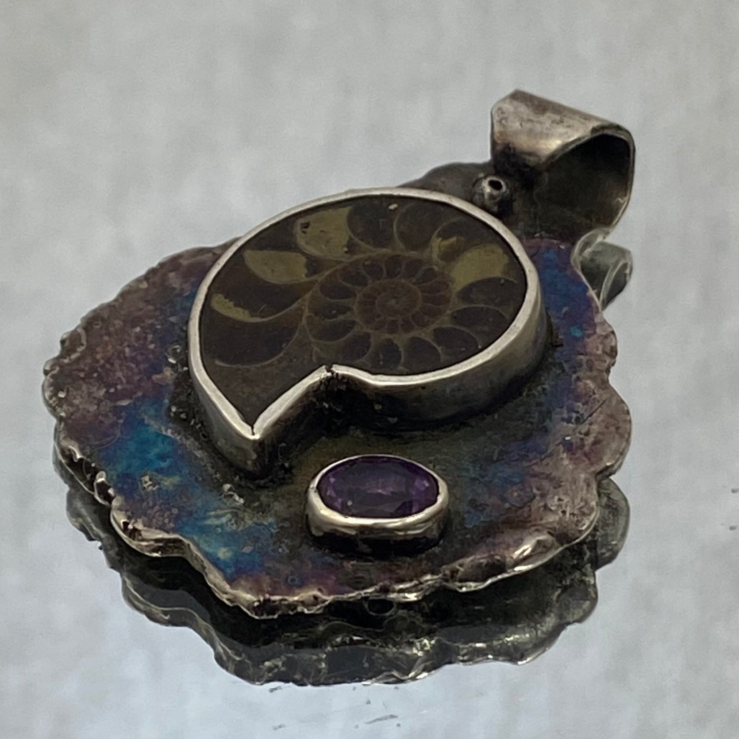 Dije de Plata Fina 950 de Colección Limitada, diseño de hoja pavonada. El plano general muestra la joya sobre fondo reflectante con una ramita decorativa con una pequeña flor blanca (Apartadó).