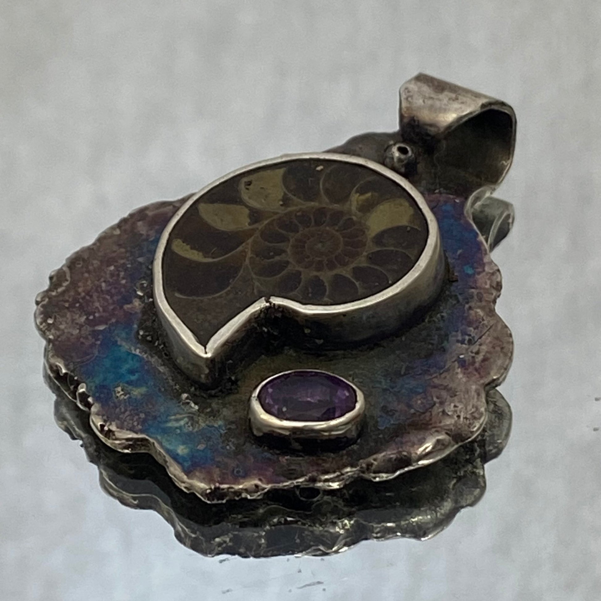 Dije de Plata Fina 950 de Colección Limitada, diseño de hoja pavonada. El plano general muestra la joya sobre fondo reflectante con una ramita decorativa con una pequeña flor blanca (Apartadó).