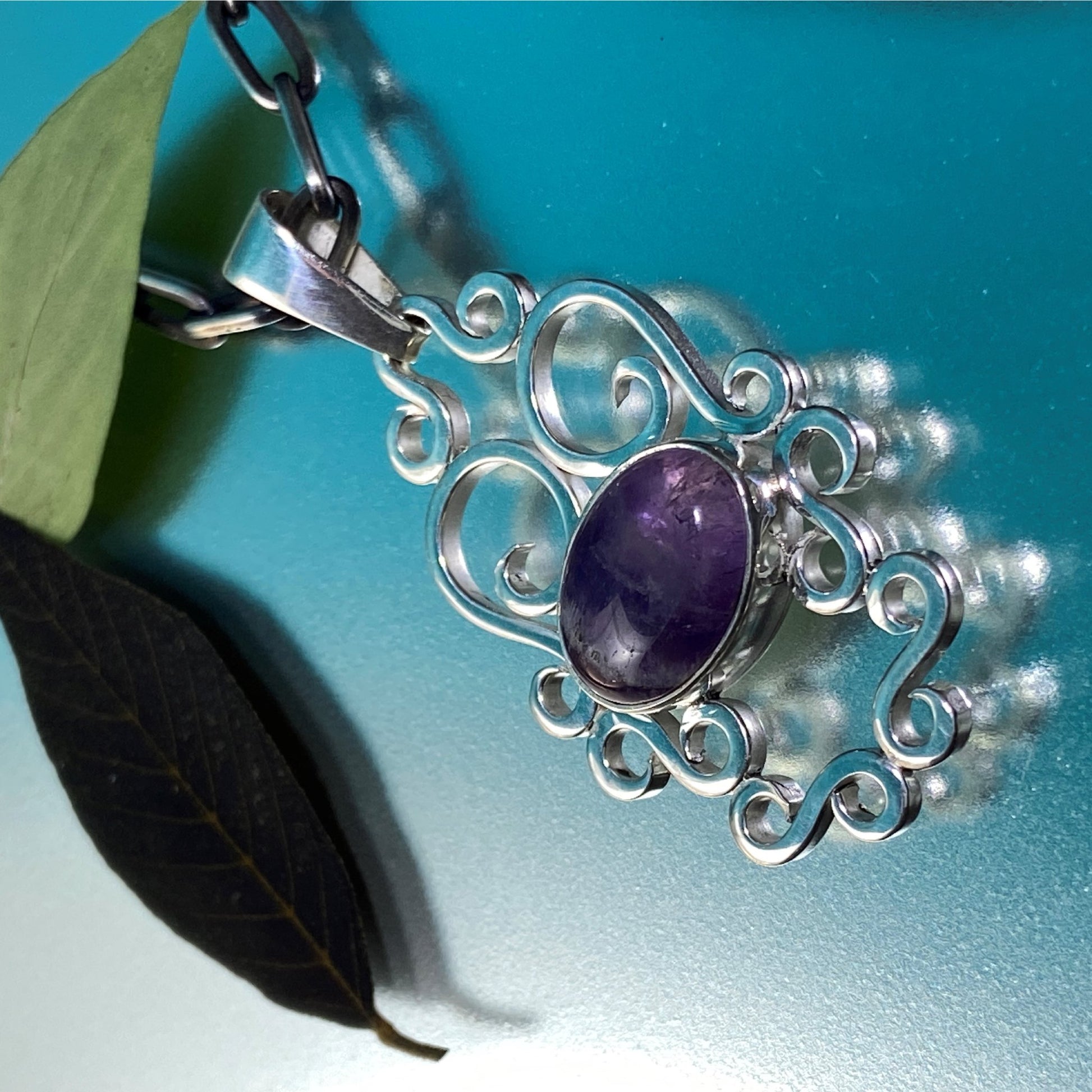 Dije de Plata Fina 950, Handmade Jewelry (Joyería Hecha a Mano). Colgante con Amatista púrpura en engaste de volutas, enmarcado por hojas frescas y oscuras sobre fondo azul (Bogotá).