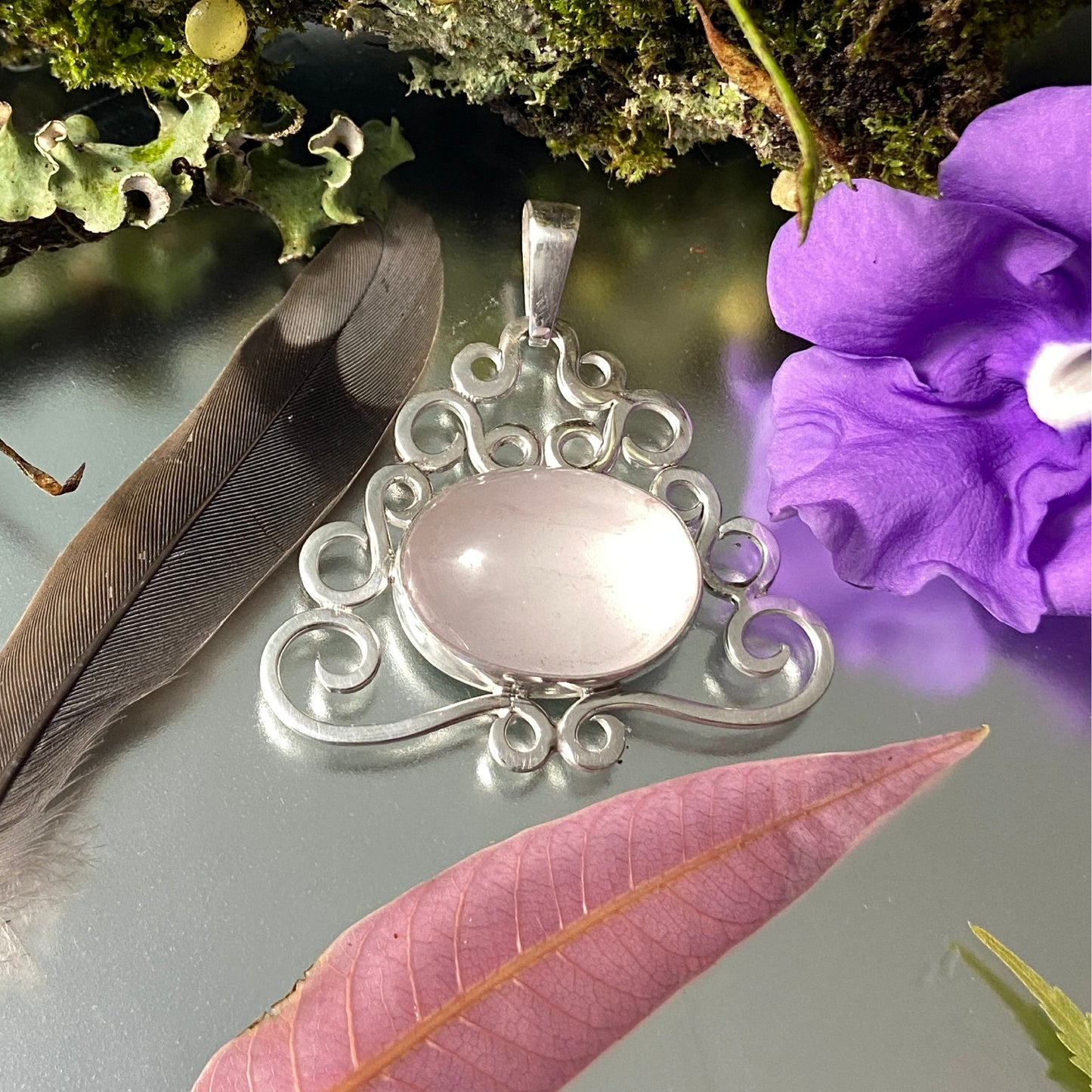 Colombian Jewelry. Dije de Cuarzo Rosado en marco de volutas. Decoración con flor púrpura intensa y pluma, destacando el acabado de la plata sobre fondo reflectante.