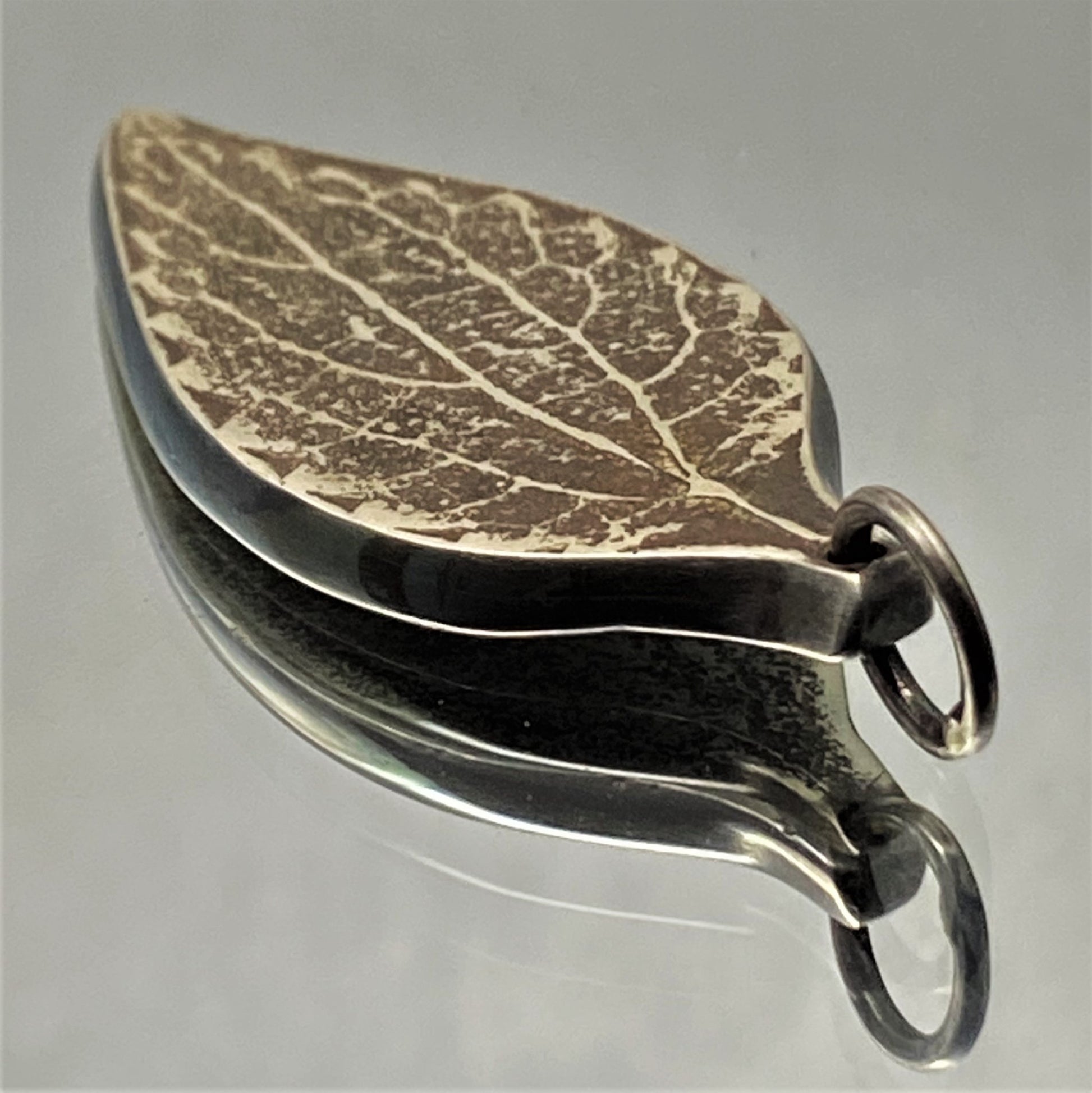 Dije de hoja de Plata Fina 950 de Colombia. Primer plano del colgante oxidado con textura de vena, pieza original con reflejo sobre superficie clara.