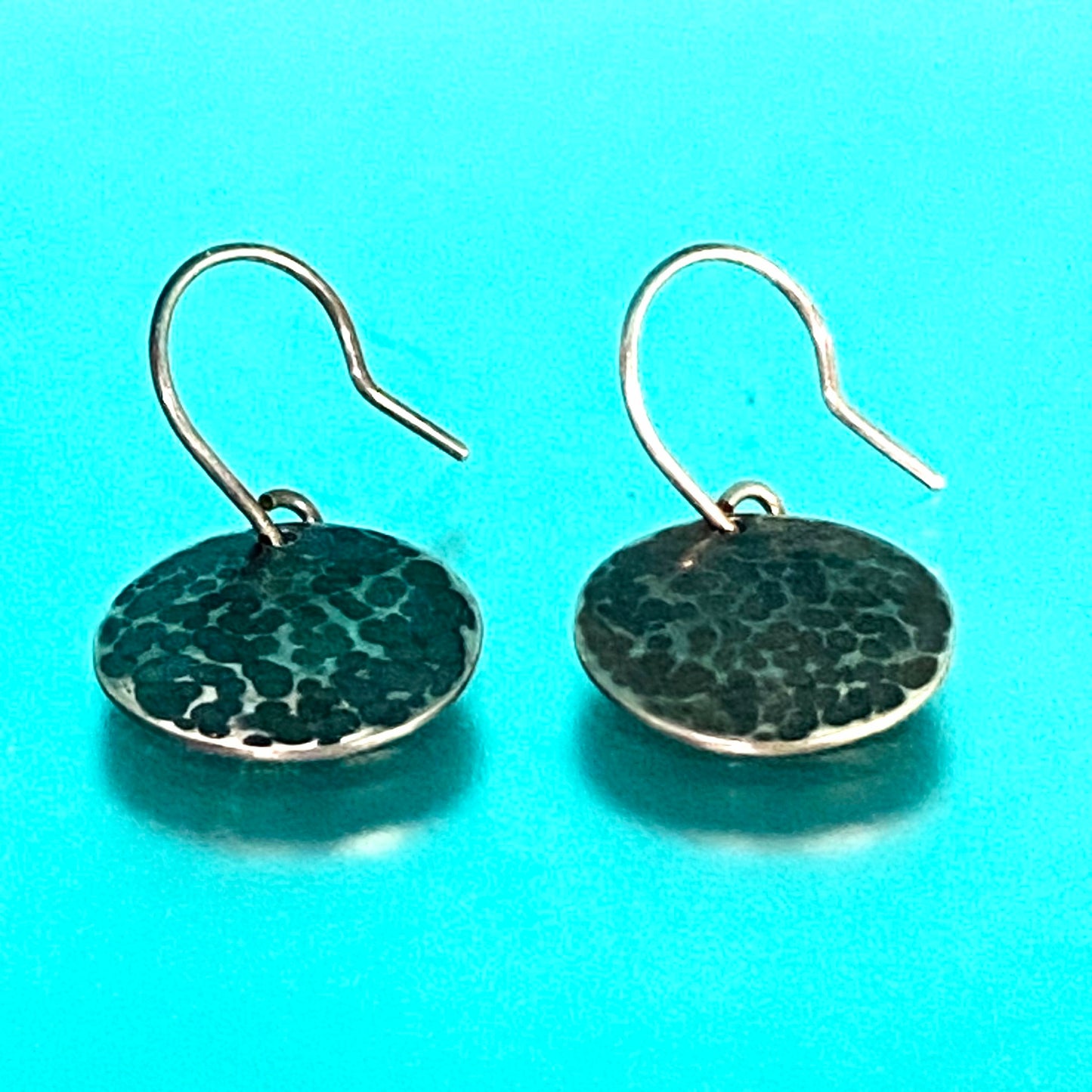 Par de Aretes de Plata Fina 950 (Martillado y Pavonado) con Joyas con Historia, vistos sobre una superficie turquesa brillante. Esta foto especial utiliza un color de fondo diferente al habitual.