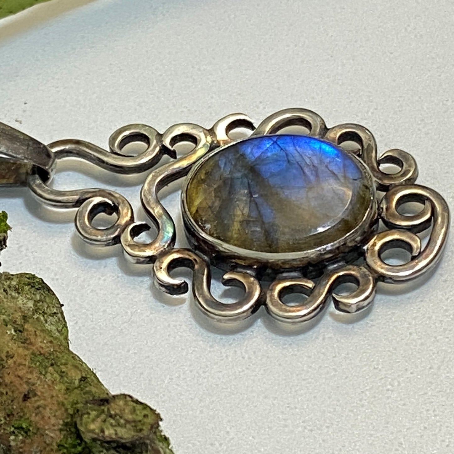 Dije de Plata Fina 950, Joyería Artesanal. Primer plano lateral horizontal de la Labradorita ovalada (destello azul), con engaste de volutas sobre fondo blanco, junto a un elemento natural con musgo.